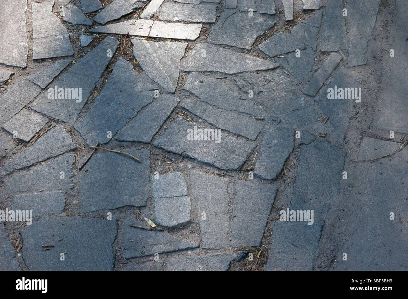 Old stone-paved path made of irregular granite slabs. The surface is uneven, with debris between the stones and small depressions from wear and age. Stock Photo