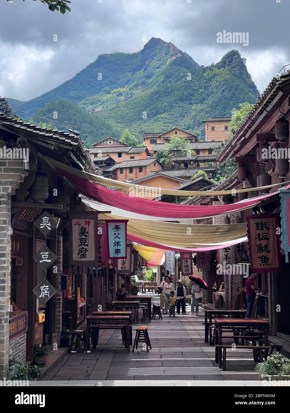 China — traditional old-town alley lined with wooden houses and Chinese signboards, colorful canopy banners, and a lush mountain rising behind. - Smartphone Captured Stock Image