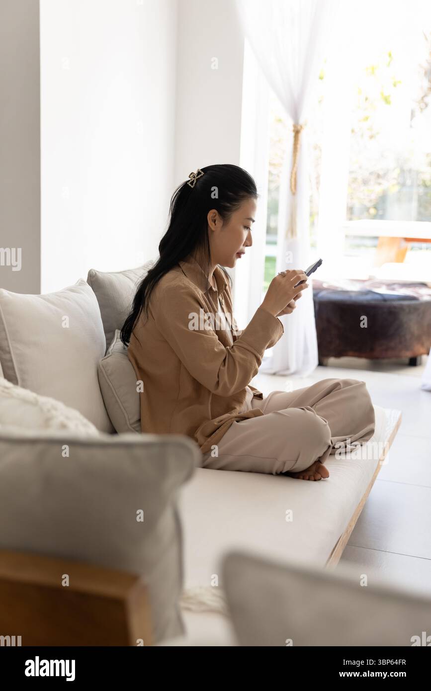 Engrossed Asian woman sits on sofa with smartphone Stock Photo
