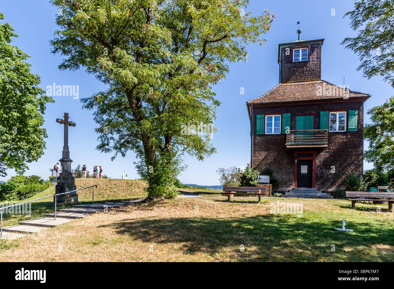 The Hochwarthaus was built in 1839 by Privy Councillor Eugen von Seyfried as a picturesque tea house and sits enthroned on the highest point of the island, about 40 metres above Lake Constance, with a panoramic view over Untersee, vegetable fields and the Rhine. Today it serves as a ceramics studio and also as a tea room again. On the island of Reichenau in Baden-Württemberg, visitors enjoy the view from Hochwart. In the foreground are a striking stone cross and a large tree, next to which is the Hochwart art gallery, which also serves as a viewing tower. The scene shows a sunny day in the reg Stock Photo