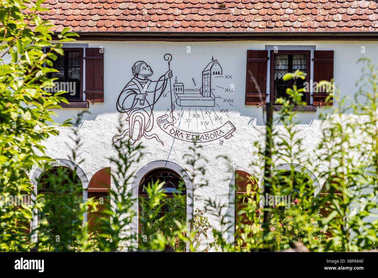 A mural on a house on Haitostraße in Mittelzell shows a Benedictine monk holding a crozier in front of a church, with the inscription "Ora et Labora" (pray and work) beneath it. The depiction possibly refers to Abbot Hatto III, who led Reichenau Abbey from 888 to 913 and later became Archbishop of Mainz. A sundial with Roman numerals is worked into the mural. Haitostraße, Reichenau, Baden-Württemberg, Germany. Stock Photo
