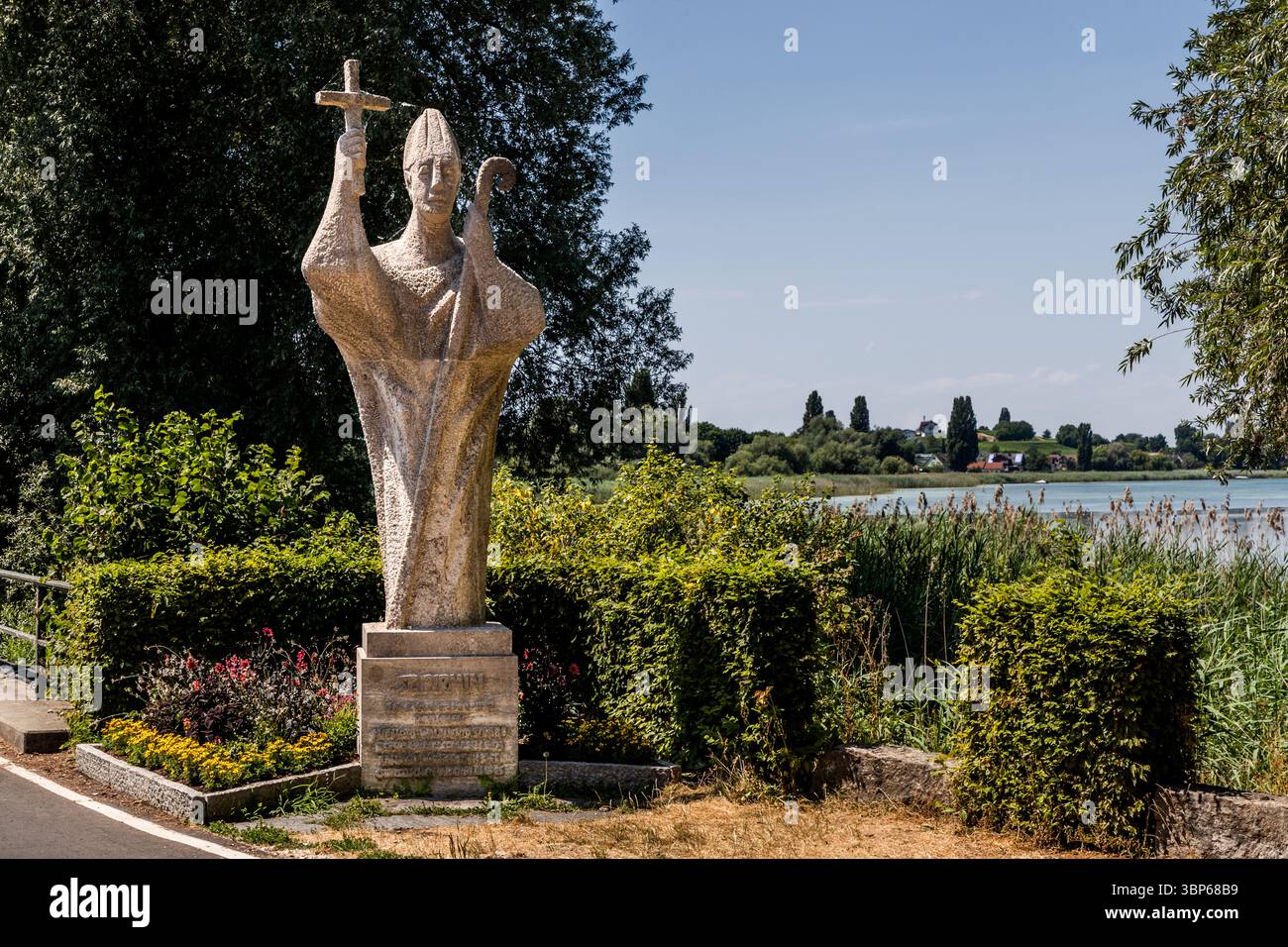 A larger-than-life stone statue of Bishop Pirmin stands at the Bruckgraben, by the bridge linking the mainland to the island of Reichenau. It commemorates his arrival in the year 724, when he founded the monastery. Pirmin is shown holding a cross and a crozier, looking out over Lake Constance. Reichenau, Baden-Württemberg, Germany. Stock Photo