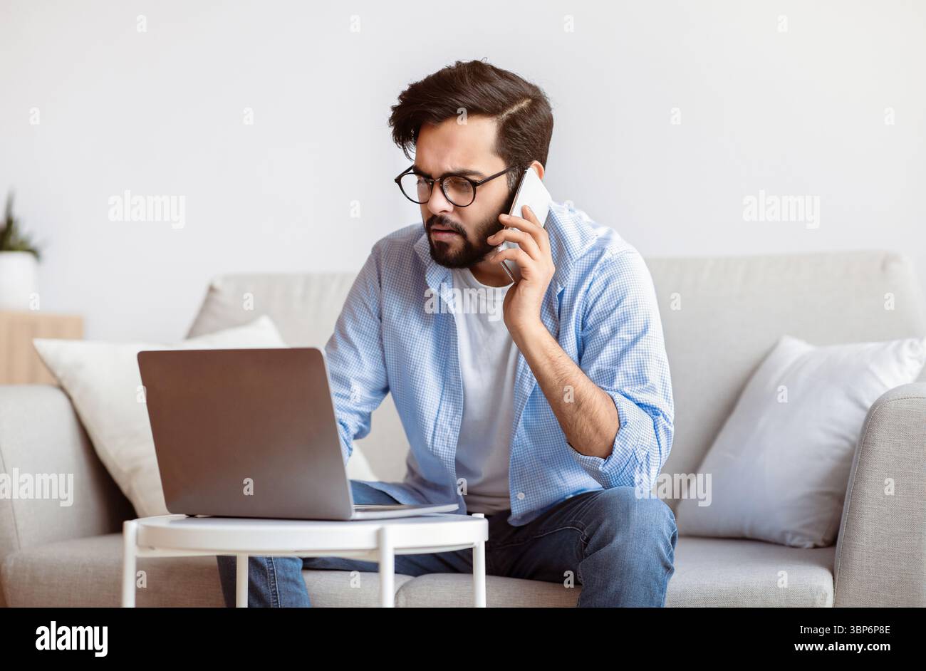 Busy Eastern Freelancer Guy Talking On Phone And Using Laptop At Home Stock Photo