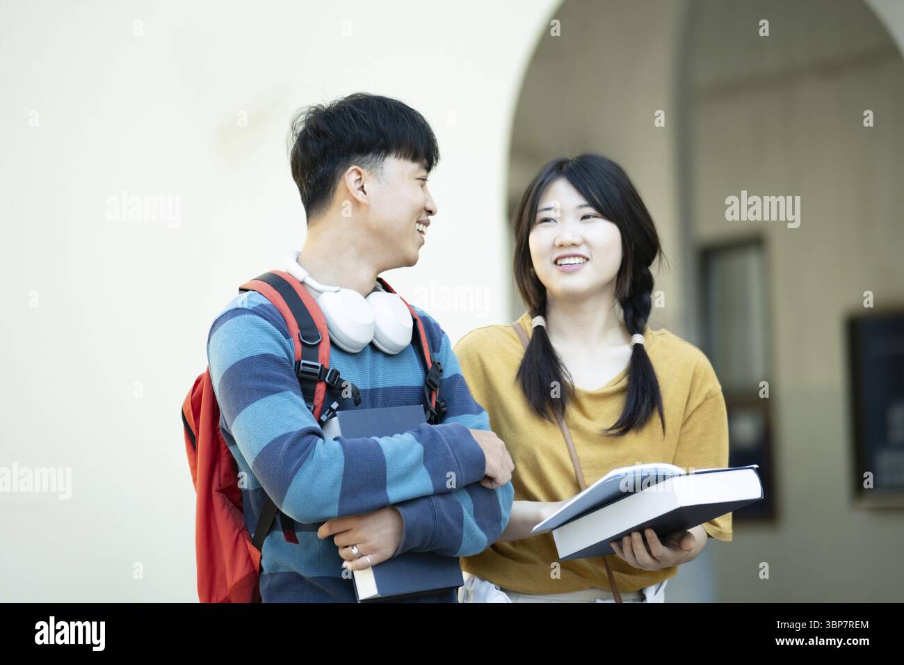 Two college students smiling and discussing their studies while holding books, with a campus building in the background and other students studying ne Stock Photo