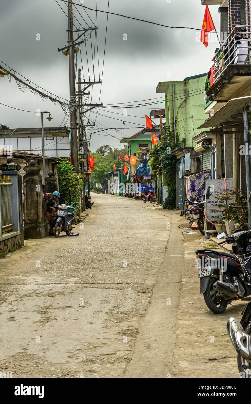 The photograph captures a narrow, unpaved street in what appears to be a Vietnamese village. The street is dusty and runs between closely packed house Stock Photo