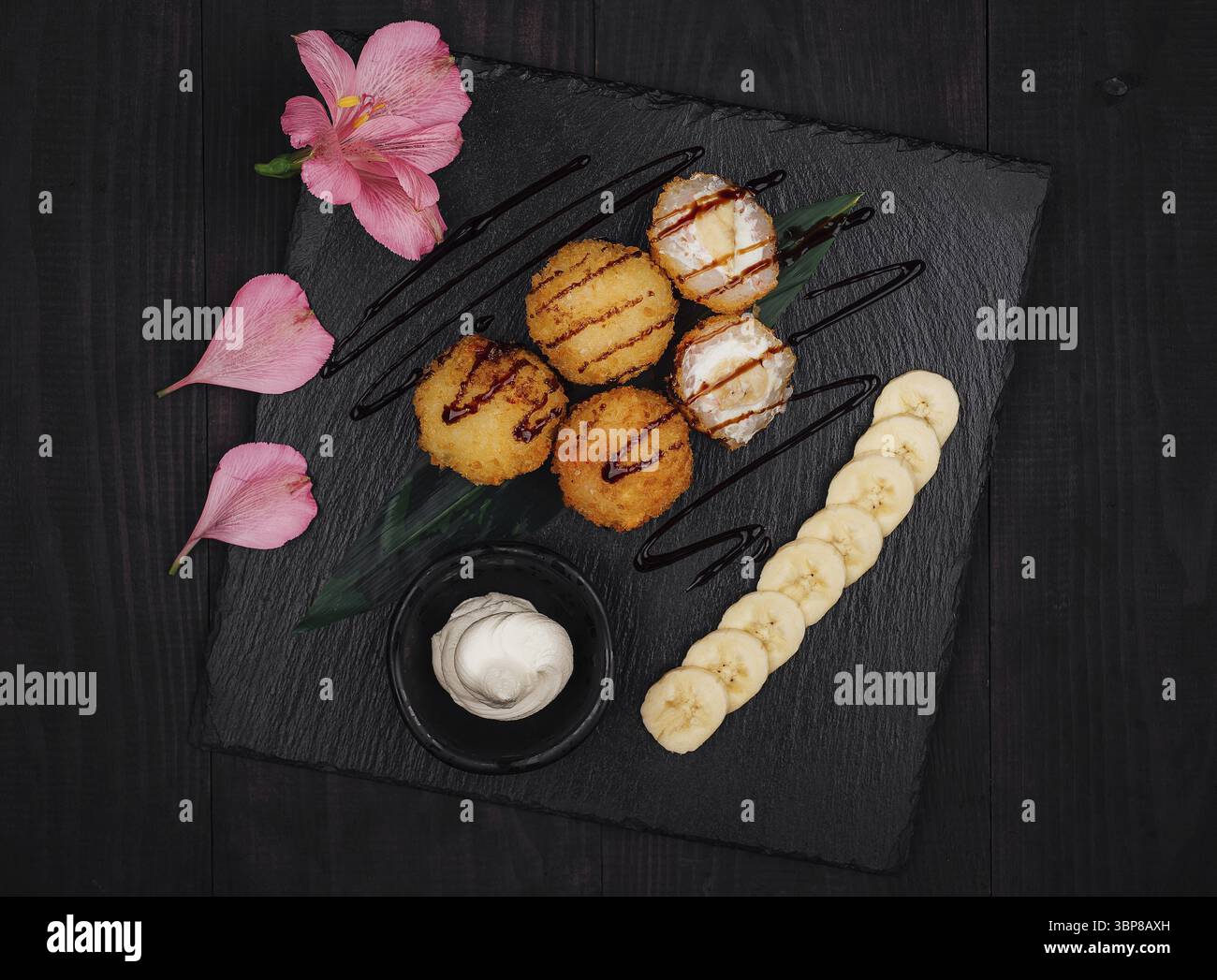 Food, Deep fried banana and cream cheese sushi balls being served on a black stone platter with chocolate syrup and sliced bananas Stock Photo