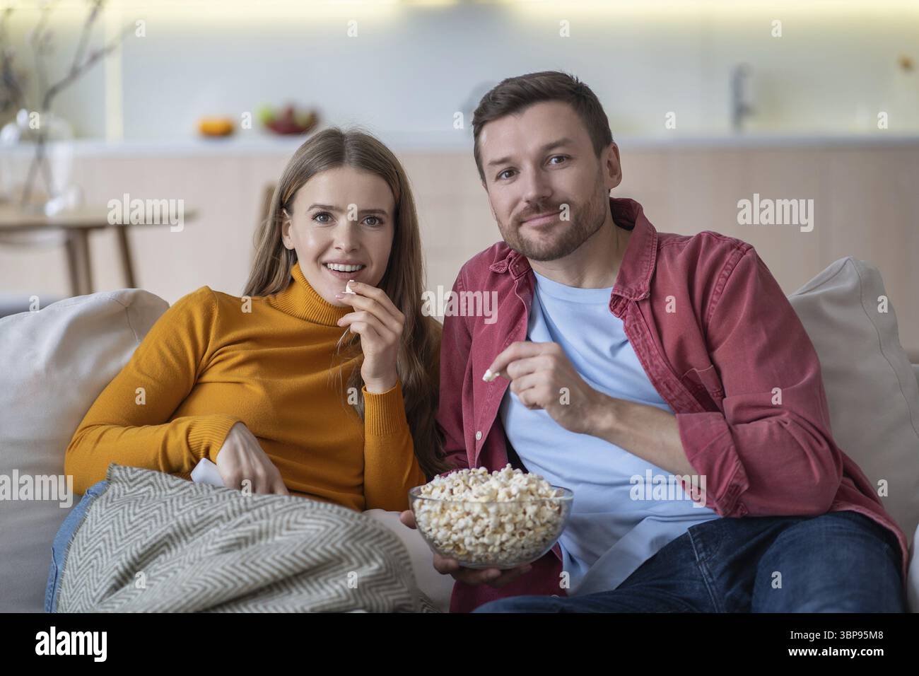 Pleased boyfriend and girlfriend enjoying watching movie while sitting comfortably on couch at home and eating tasty popcorn. Happy interested loving Stock Photo