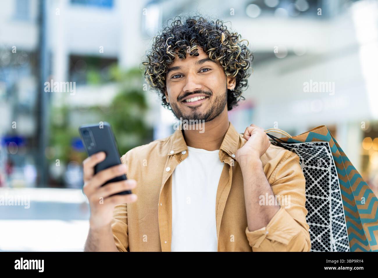 A smiling man holds his phone while carrying shopping bags, enjoying a day of shopping and using technology. Stock Photo
