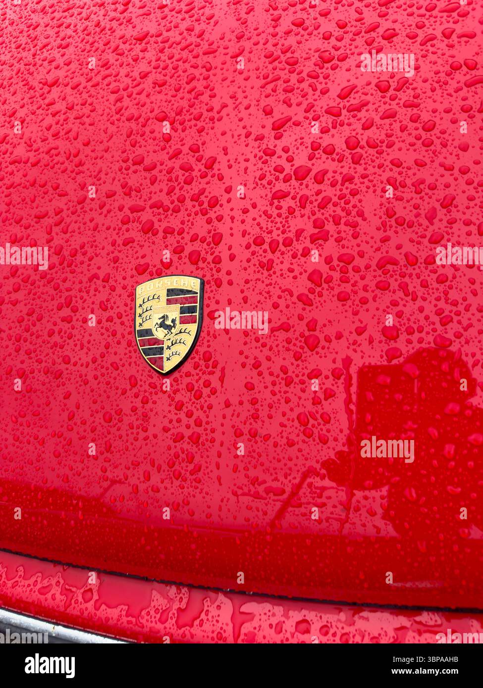 Close-up of a wet red Porsche 911 car hood or bonnet featuring a Porsche logo covered in raindrops reflecting light - Smartphone Captured Stock Image
