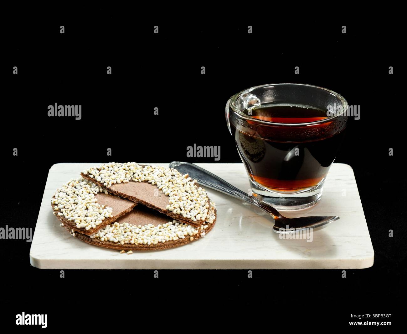 Glass cup of hot coffee paired with a traditional Mexican-style chocolate and puffed amaranth cookie on a white marble board with a spoon Stock Photo