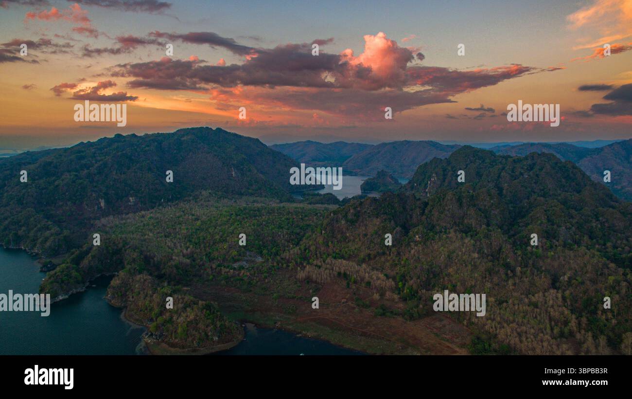 A breathtaking aerial view captures the soft glow of sunrise casting golden reflections over the tranquil waters of Vajiralongkorn Dam. Dotted with lu Stock Photo