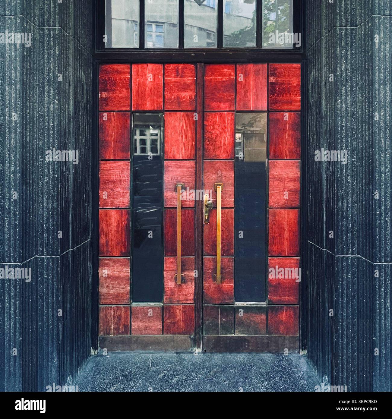 Modernist red tiled double doors with brass handles, framed by grooved dark stone — a bold architectural entrance in central Kraków, Poland. - Smartphone Captured Stock Image