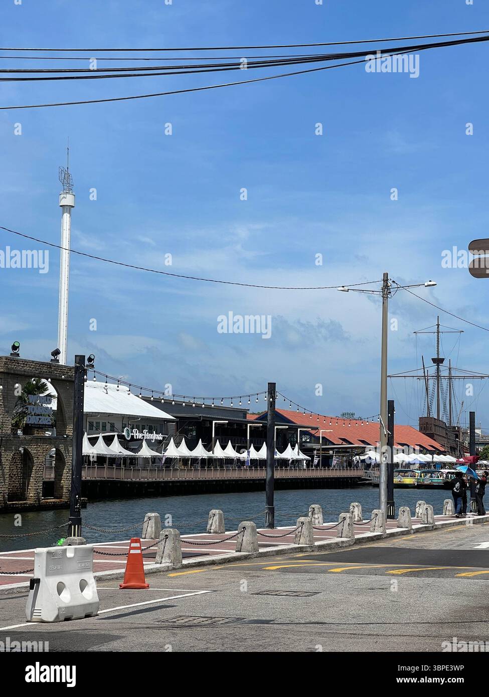 Malacca, Malaysia - June 21, 2025: View of the Malacca River waterfront with shop buildings, riverside promenade, tourist boats, and pedestrians. - Smartphone Captured Stock Image