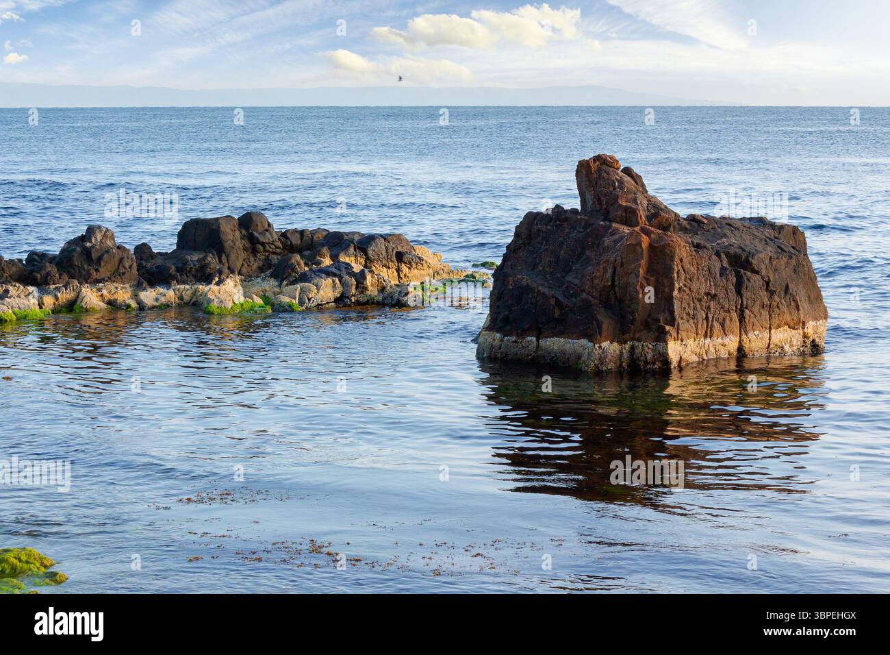 seascape with rock in the water. textured boulder reflecting calm sea surface in morning light. vacation season in summer. sunny weather. fluffy cloud Stock Photo