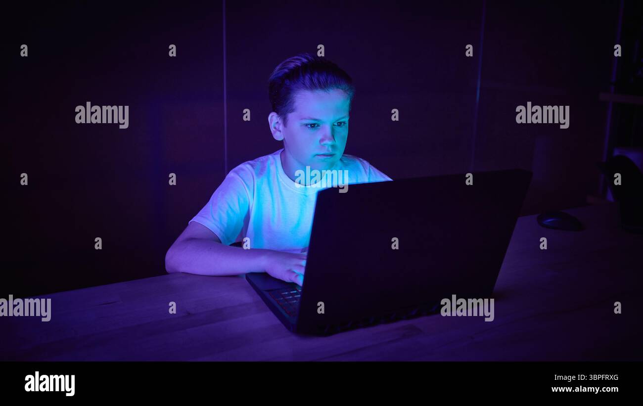 Focused boy using laptop in dark room with blue lighting Stock Photo