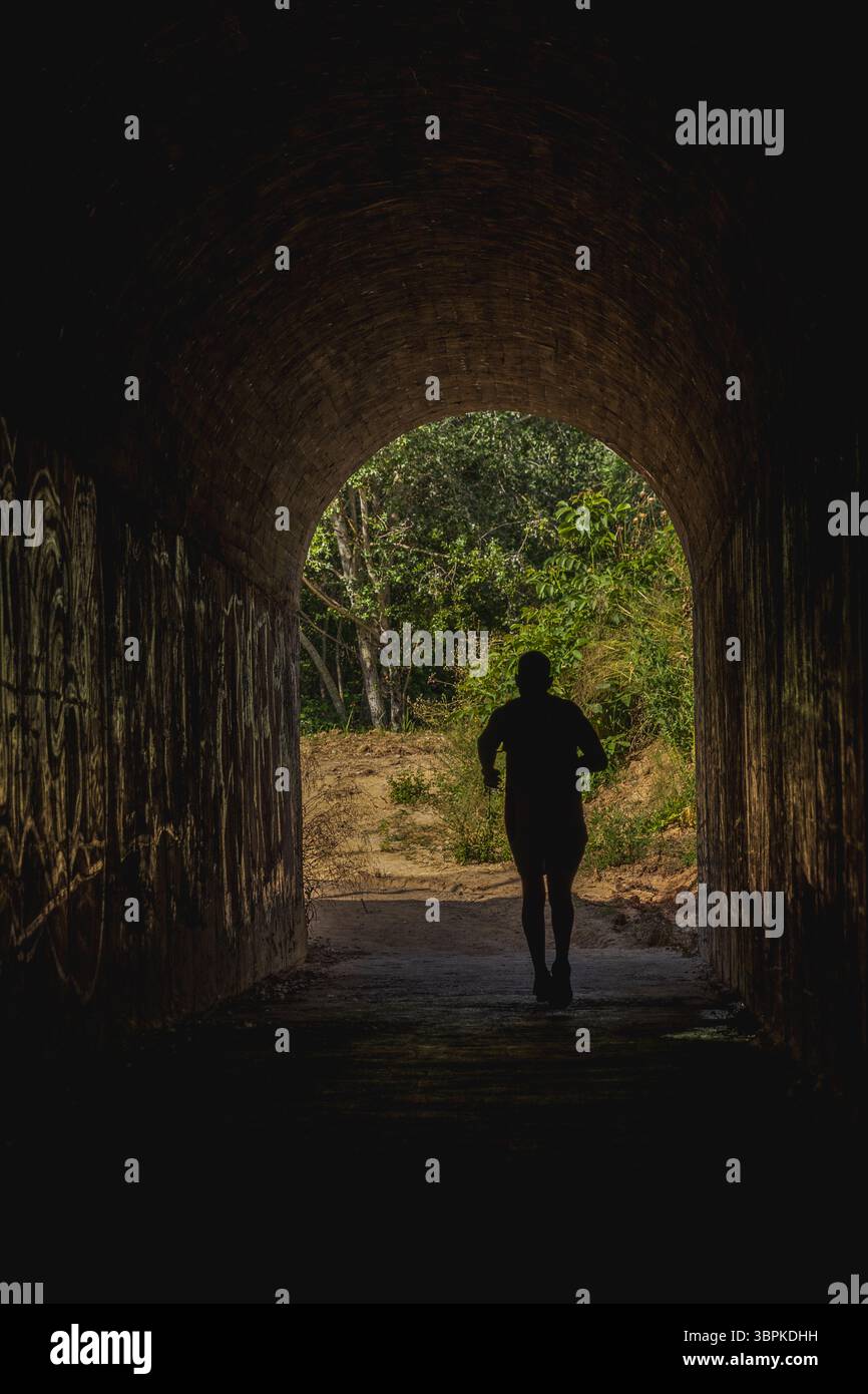 Lone runner's silhouette emerges from a dark tunnel, framed by lush greenery outside. The scene captures the essence of solitude and determination in Stock Photo