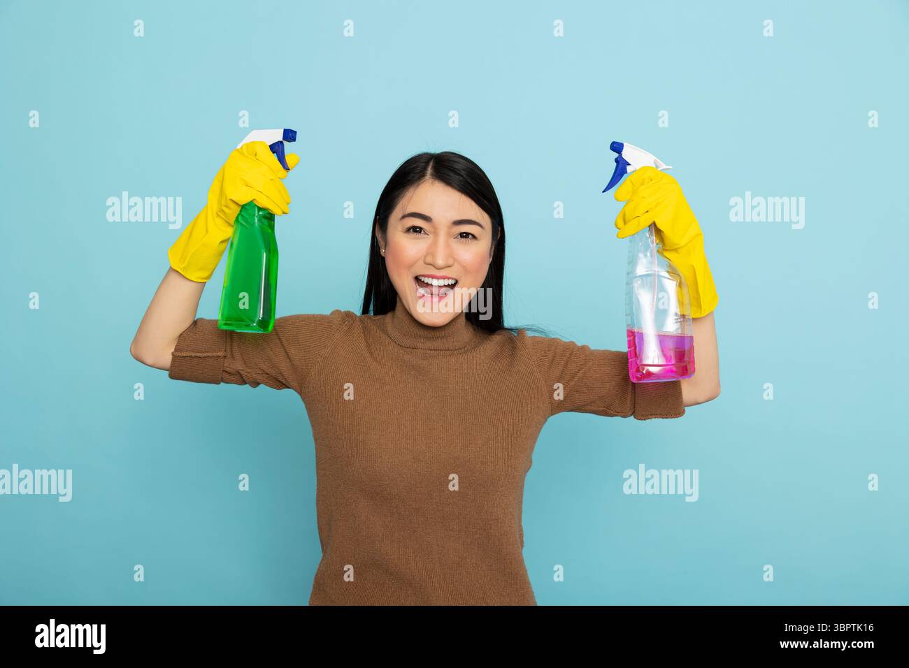 Energetic asian housekeeper in rubber gloves holding colorful cleaning sprays and flexing arms, promoting cleanliness with big smile. Enthusiastic female cleaner with pink and green liquid solutions. Stock Photo
