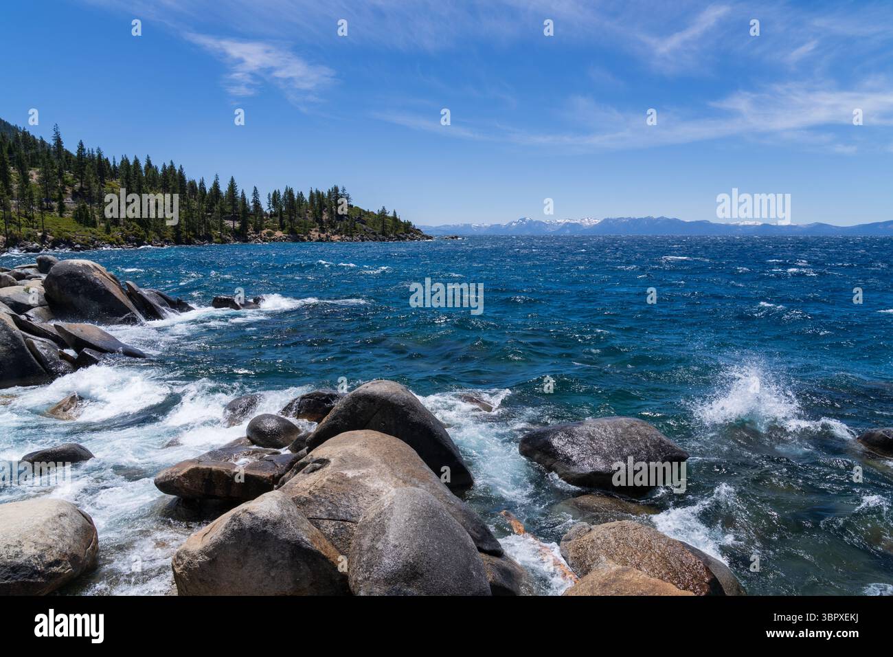 Lake Tahoe, California, USA, high winds creating waves splashing against the rocks, blue sky and distant snowy mountains Stock Photo