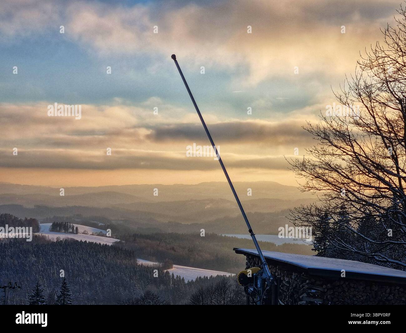 A tilted pole creates a stark, abstract silhouette against a moody, dramatic orange sunset sky over a rolling winter landscape in the Czech mountains. - Smartphone Captured Stock Image