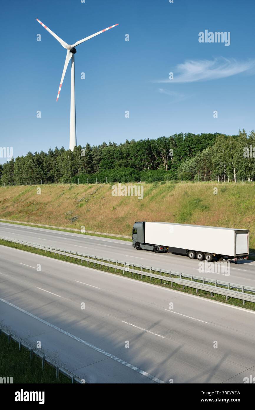 Long haul truck driving on highway with wind turbine in background, representing modern logistics, transportation infrastructure, and renewable energy Stock Photo