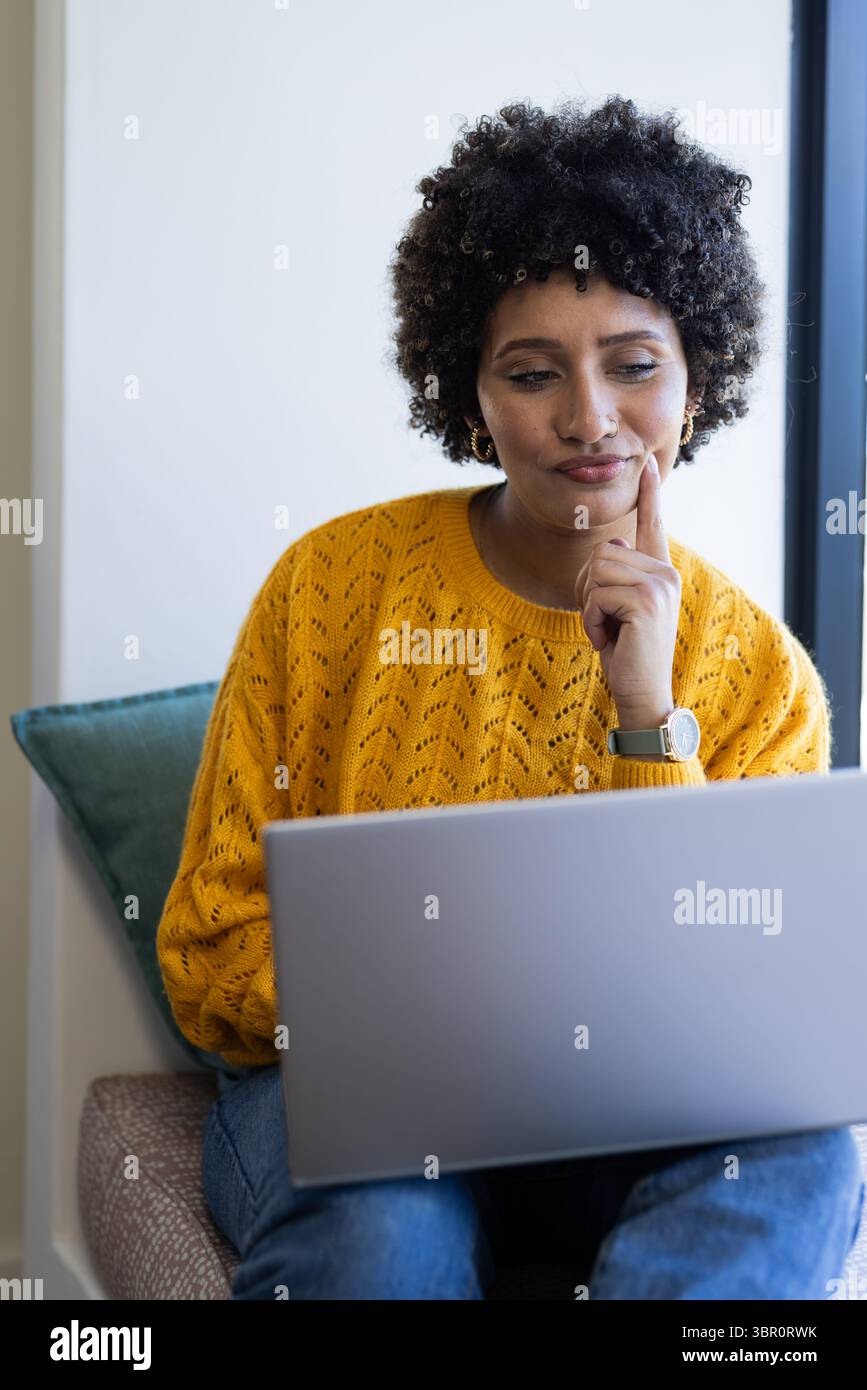 African American woman in yellow sweater sitting on teal cushion by window typing on silver laptop. Casual, workspace, modern, comfort, concentration, Stock Photo