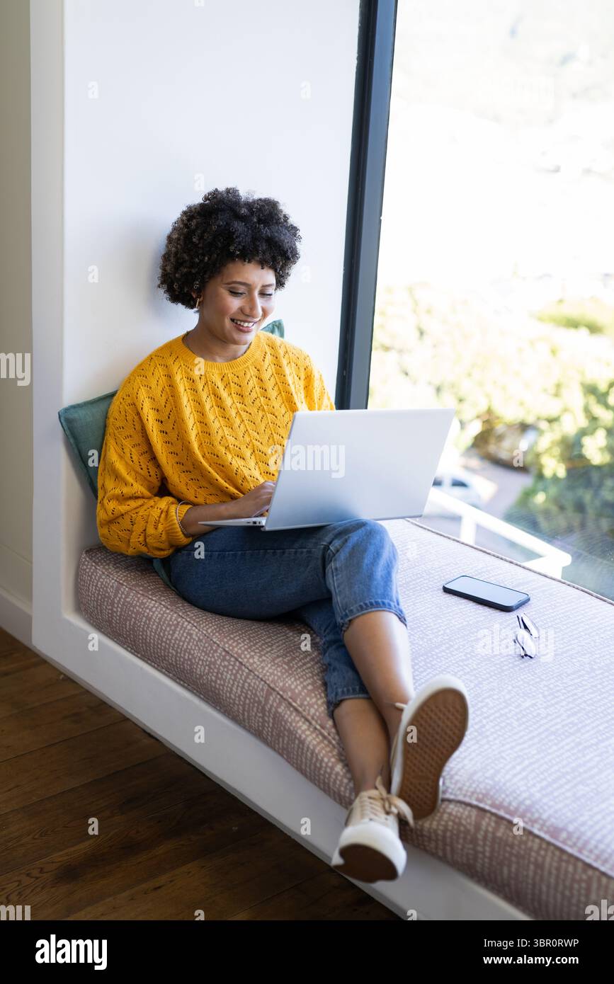 African American woman sitting on window seat in reading nook, using laptop with smartphone. Cozy, leisure, workspace, digital, nature, lifestyle, mod Stock Photo