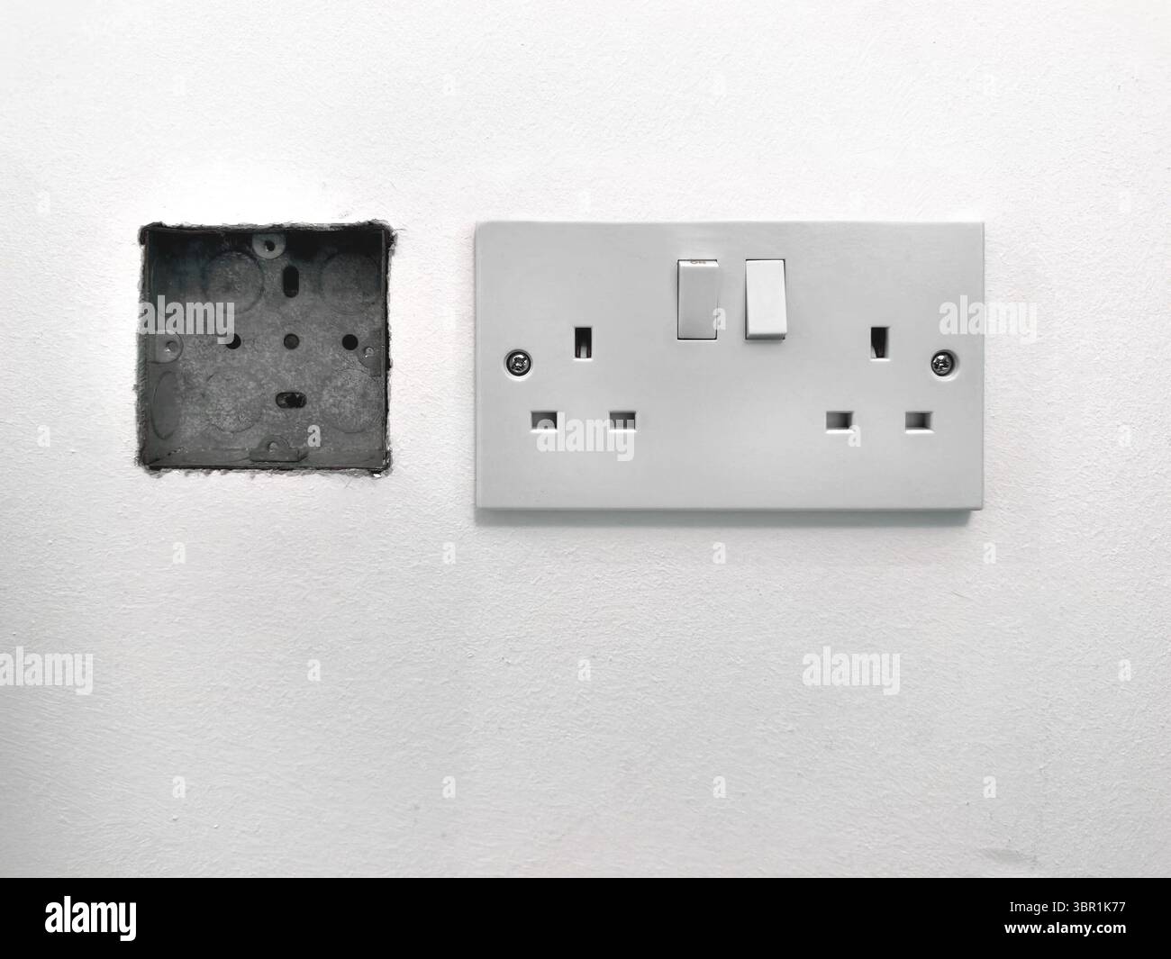 Close-up view of a UK double electrical outlet with switches next to an empty electrical wall cavity box on a plain white wall - Smartphone Captured Stock Image