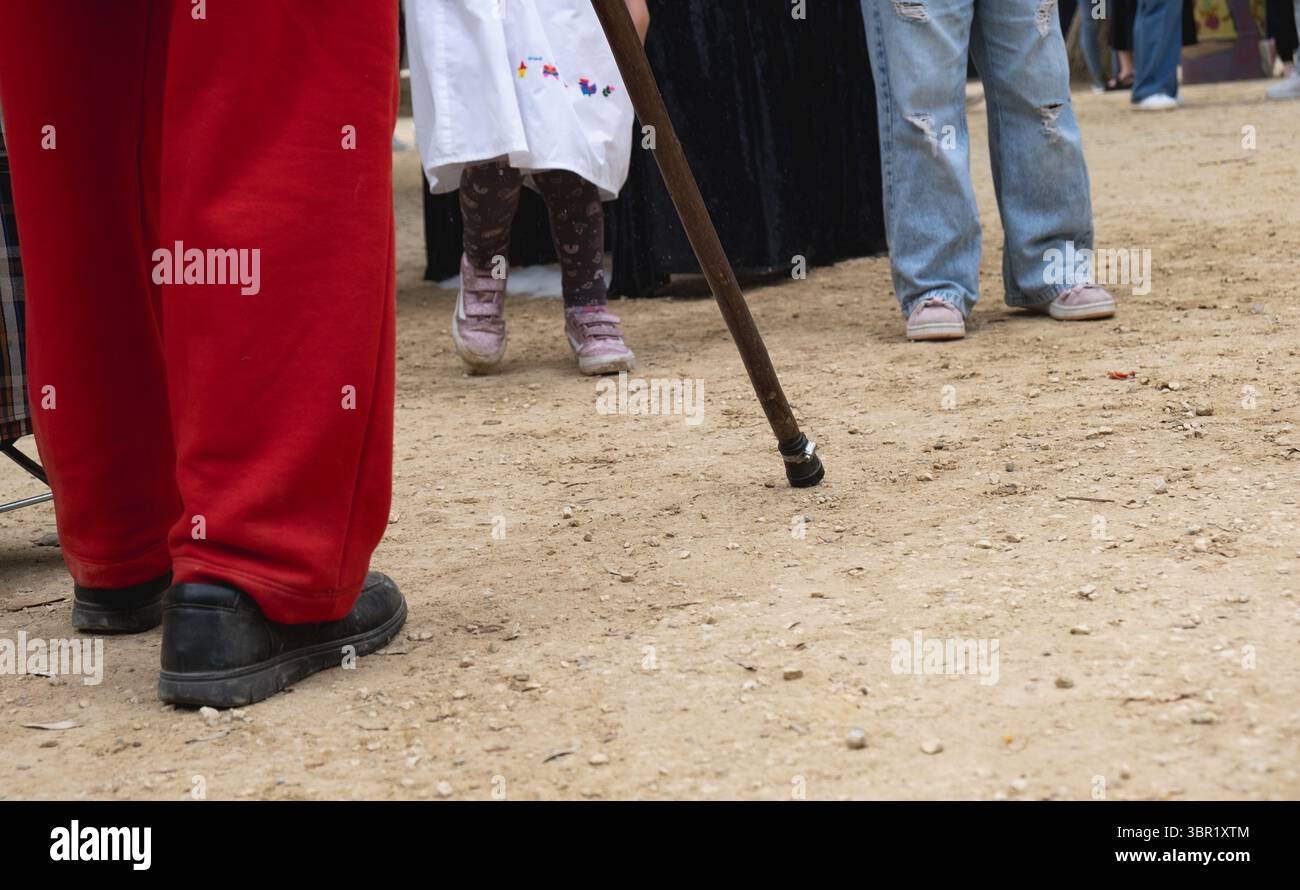 Street performer wearing red pants and black shoes standing still with walking stick on dirt ground, audience and child in background Stock Photo