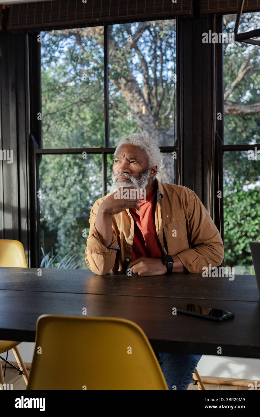 Senior African American man sitting at home dining table under bamboo blinds gazing with smartphone. Contemplation, introspection, tranquility, modern Stock Photo