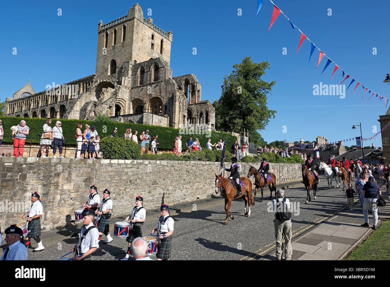 Jedburgh, UK. 11th July, 2025. Jethart Callant's Festival 2025, Passing the ruins of Jedburgh Abbey, Jethart Callant 2025 Lewis Ronald Turnbull carries the Burgh Standard at the start of Festival Day, in the distance Right Hand: Lewis Stewart, Left Hand: William Wardrop. Ahead of over 100 mounted supporters during Festival Day on Friday 11th July. ( Credit: Rob Gray/Alamy Live News Stock Photo