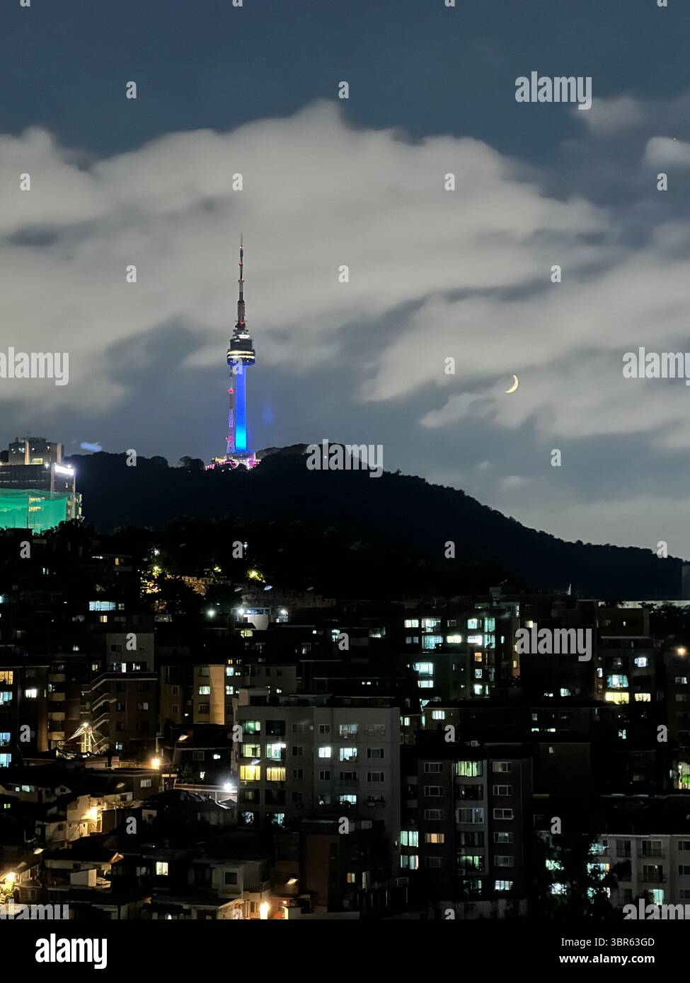 Namsan Seoul Tower illuminated in blue at dusk with dramatic clouds above Seoul skyline, South Korea, viewed from hillside neighborhood. - Smartphone Captured Stock Image