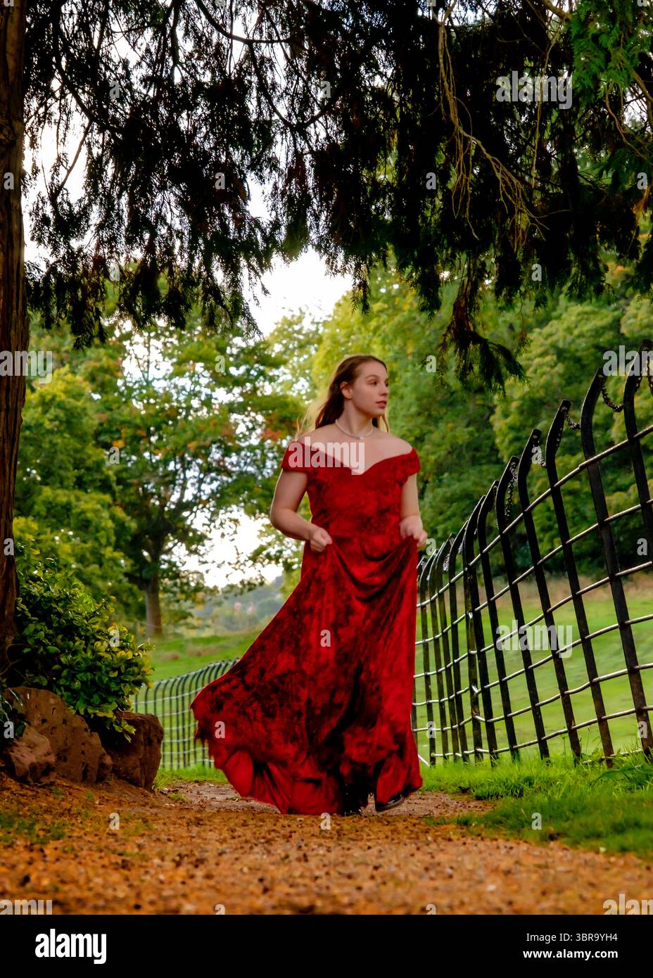 A young woman in a red dress walks along a path in a park. Stock Photo