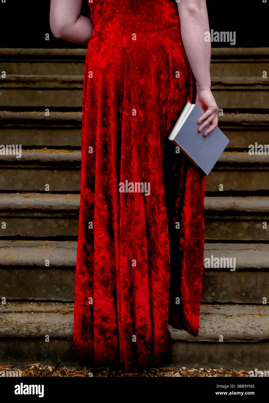 A person in a long red dress stands on a set of stairs, holding a book. Stock Photo
