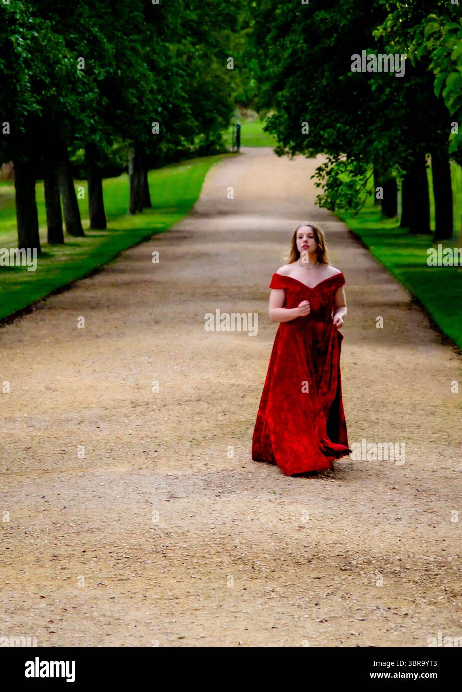 A young woman in a red dress walks down a path lined with trees. Stock Photo