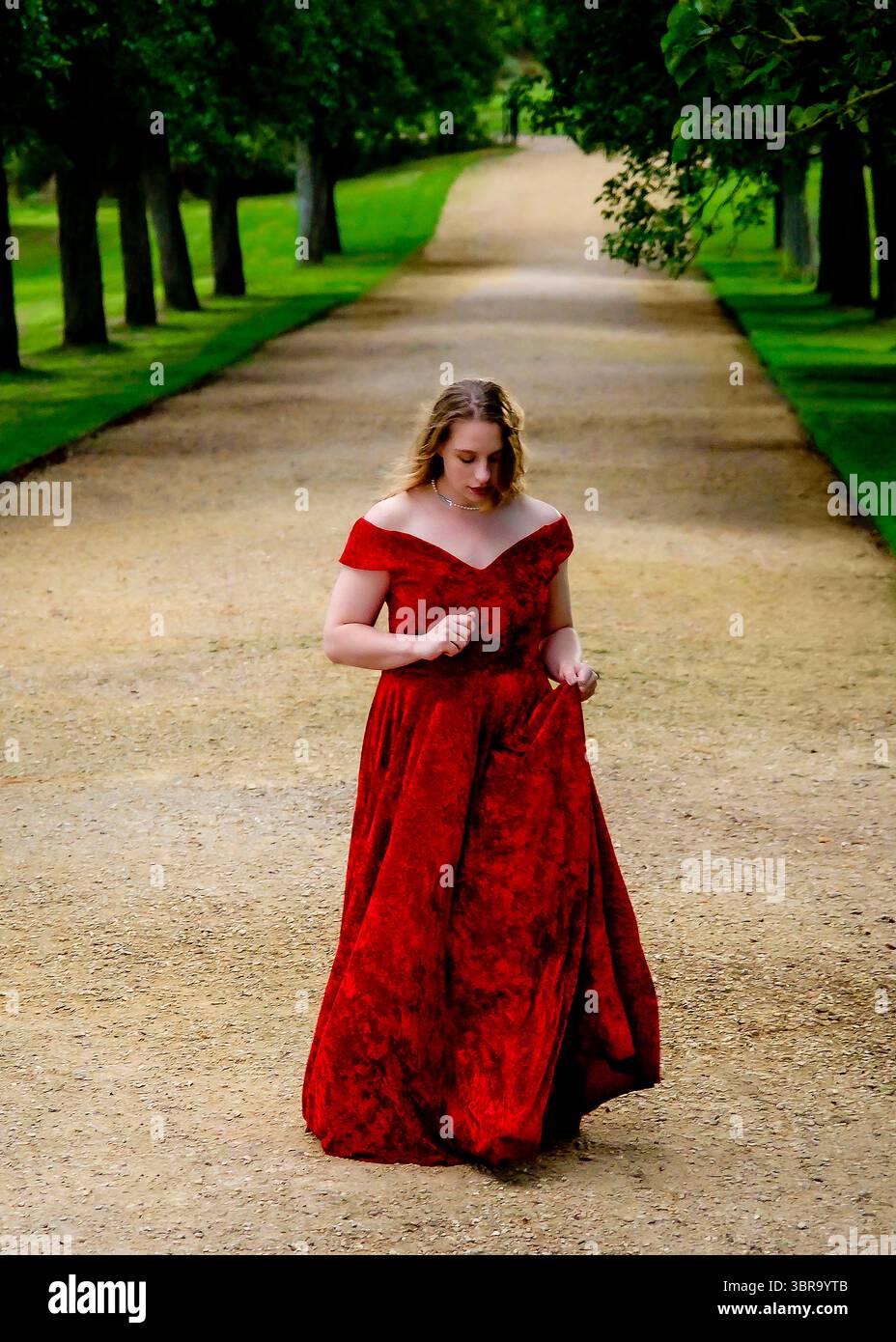 A woman in a red dress walks down a path lined with trees. Stock Photo