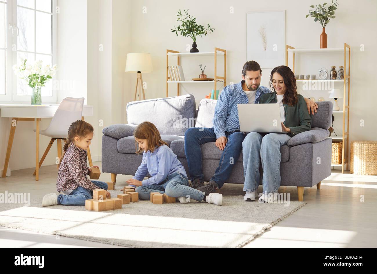 Family at home, daughter and son playing at floor with cubes, parents sit on sofa resting using pc Stock Photo