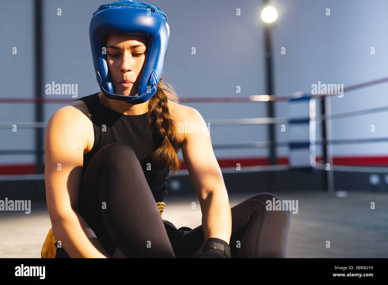 Female boxer in black gear tightening hand wraps inside boxing ring on grey mat under spotlight Stock Photo