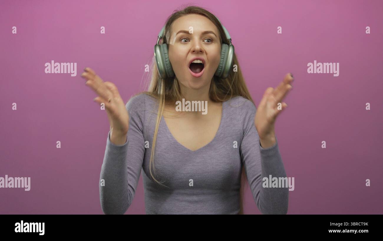 Woman enjoying music with headphones against an isolated pink background, expressing surprise and delight, wearing casual gray top, communicating happ Stock Photo