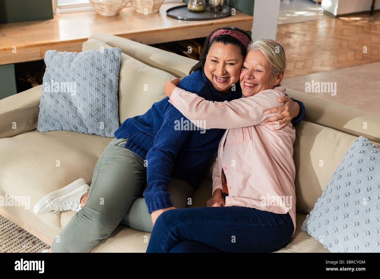 Diverse senior friends sitting on sofa at home hugging near throw pillows and console table Stock Photo