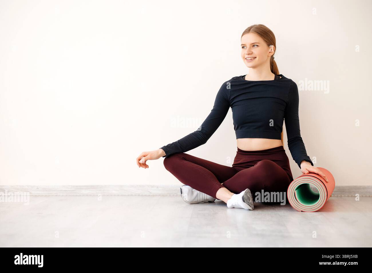 Attractive positive fit woman sitting on floor in lotus position and touching yoga mat with hand. Slim lady dressed in sport uniform looking aside and Stock Photo