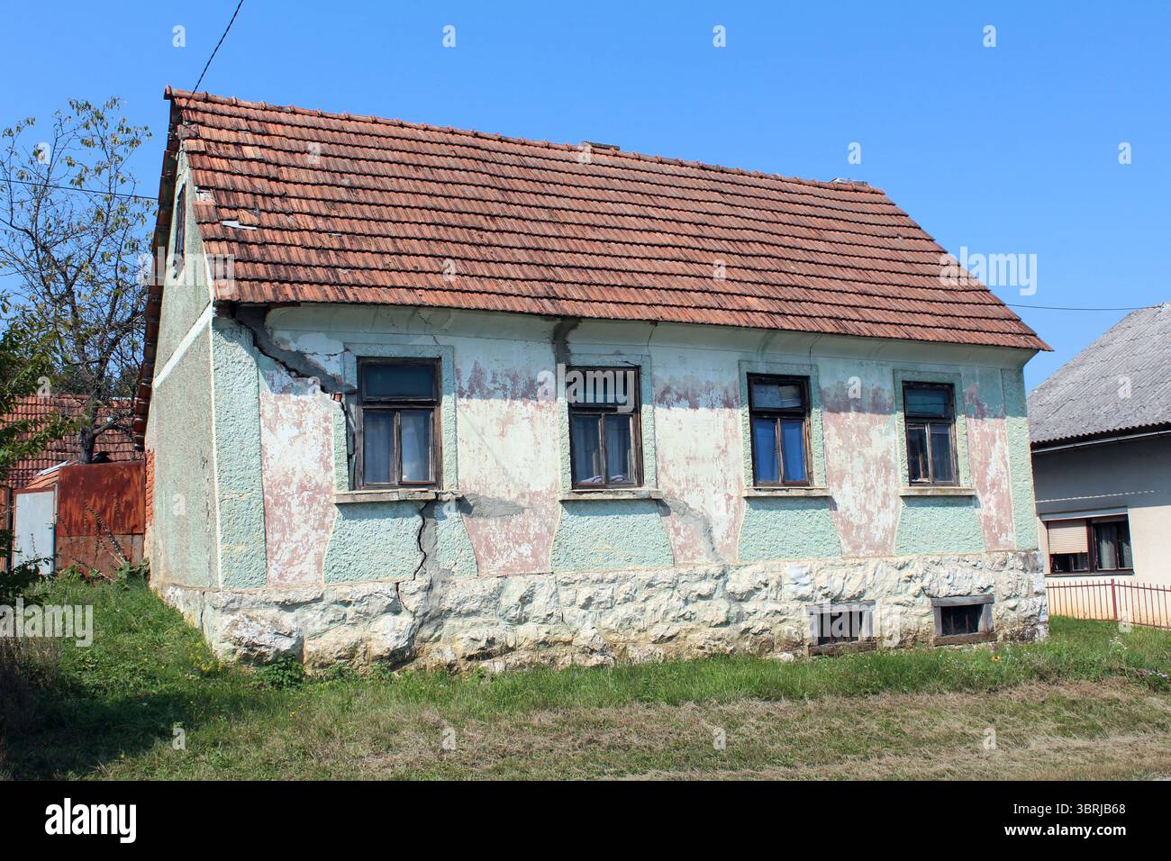 A small rural house with severe wall cracks, peeling blue-green paint ...