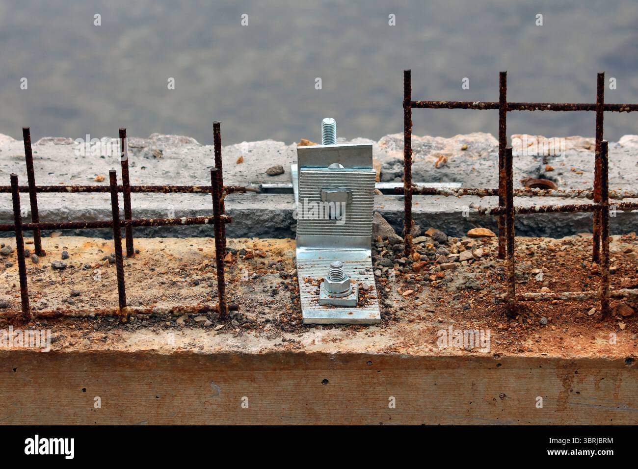 A galvanized steel anchor bracket is bolted to a concrete beam, surrounded by exposed and rusted rebar, highlighting early construction stages Stock Photo