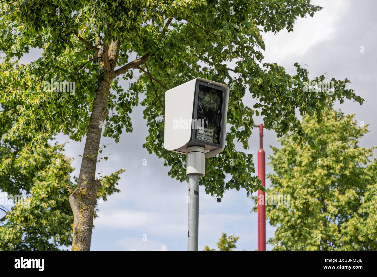 A modern white surveillance speed camera (flitspaal) mounted on a pole amidst green trees and a bright sky, ensuring safety and monitoring. Stock Photo
