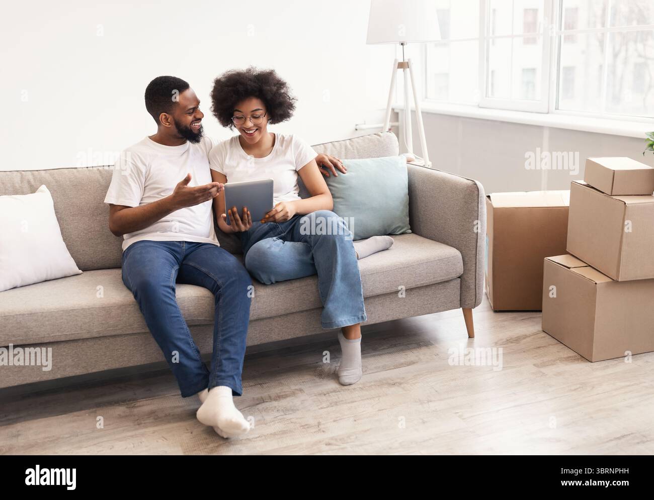 Happy Black Spouses Using Tablet Sitting Among Moving Boxes Indoor Stock Photo