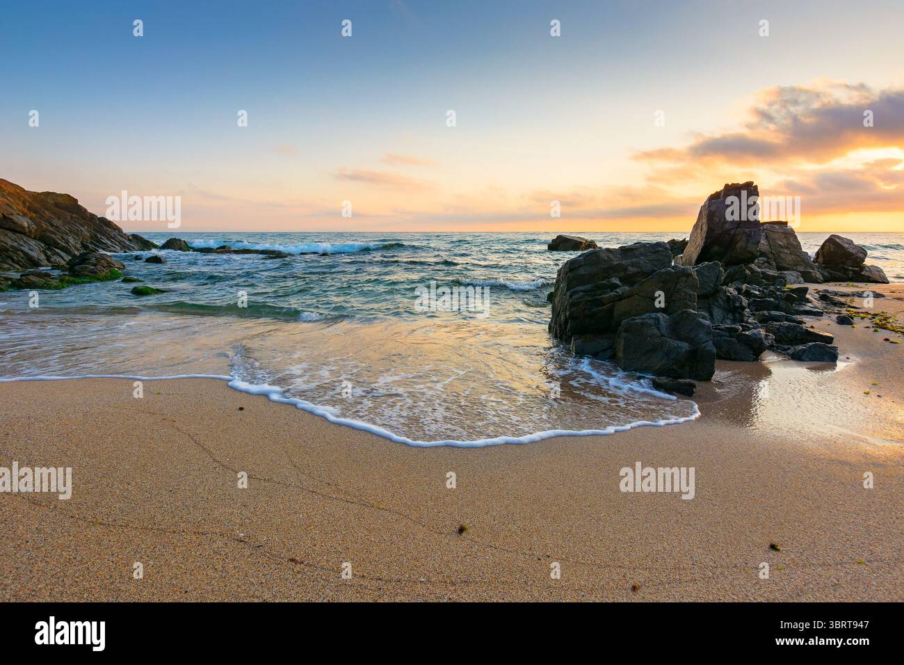 landscape by the sea at sunrise in summer. beautiful beach with rocks washed by calm waves in orange light. view to horizon under blue sky with clouds Stock Photo