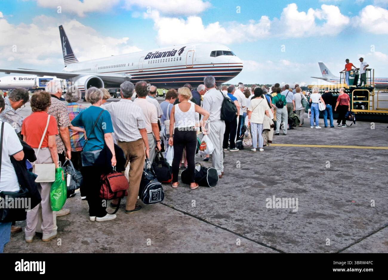 1998 Archive view of air travel Britannia Airways jet airliner combining with Thomson package holiday overlooking these returning passengers with hand luggage some connecting from 90s Caribbean cruise ship journey waiting inline wearing 90s fashions overlooked by ground crew on the airport tarmac on hot sunny day queuing up waiting to  board   fuselage of Britannia Airways Boeing jet airliner for 90s return to London Gatwick UK wearing 1990s holiday fashion at Santo Domingo AirPort a popular destination for ongoing visits to the Caribbean. It was renamed as Las Américas International in 2002 Stock Photo