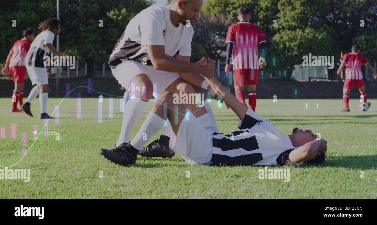 Kneeling teammate in white and black kit examining injured leg on soccer pitch, with soccer ball Stock Photo