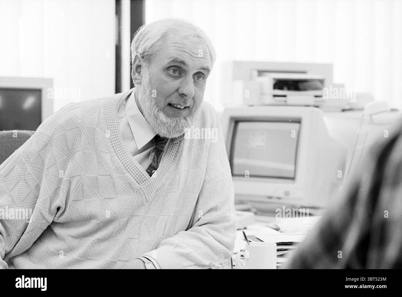 Mr. Brambach Schiphol Schiphol, Whizgle News, Dutch Desk, The Netherlands, 1950 - 2000 on 30-04-1995. The image contains these topics. In a warmly lit office setting, a gentleman with a distinguished appearance sits at a desk, engaging in conversation. His attire consists of a light-colored, knitted sweater adorned with a subtle diamond pattern, complemented by a tailored shirt and a tie that adds a hint of personality. He has a full, well-groomed beard and a thoughtful expression, enhancing his authoritative yet approachable demeanor. Around him, the workspace is organized, featuring various Stock Photo
