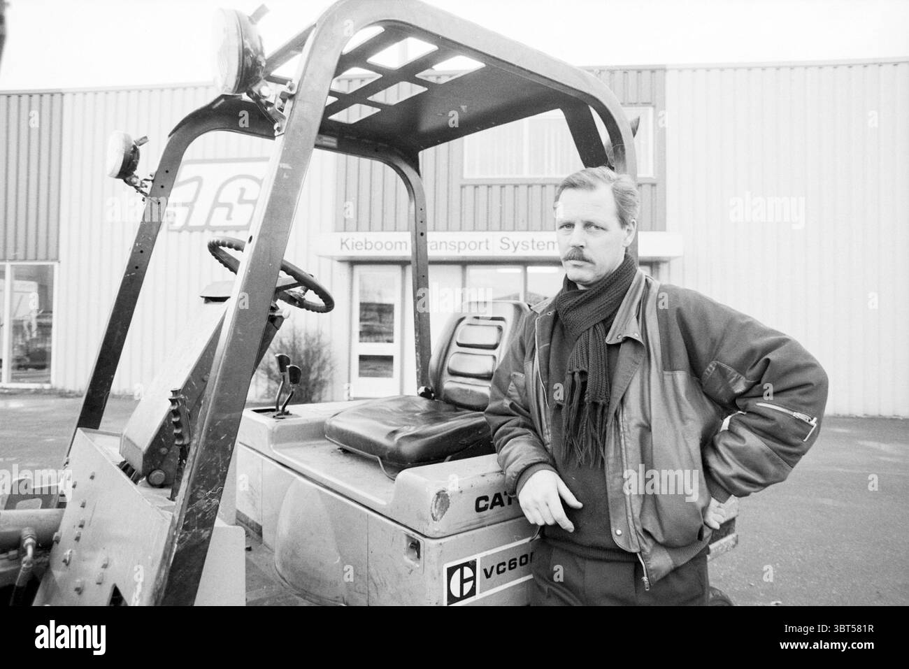 Man and forklift truck at Kieboom Transportsystemen (KTS), Whizgle News, Dutch Desk, The Netherlands, 1950 - 2000 on 11-12-1989. These are the topics in the image. The scene depicts a man standing next to a rugged, industrial vehicle. He has a serious expression, with mustache and medium-length hair, dressed in a dark-colored jacket and a thick scarf wrapped around his neck. The texture of his clothing contrasts with the smooth metal of the vehicle. The vehicle is a compact tractor with a prominent steering wheel that is somewhat weathered, hinting at its use in outdoor work environments. Its Stock Photo