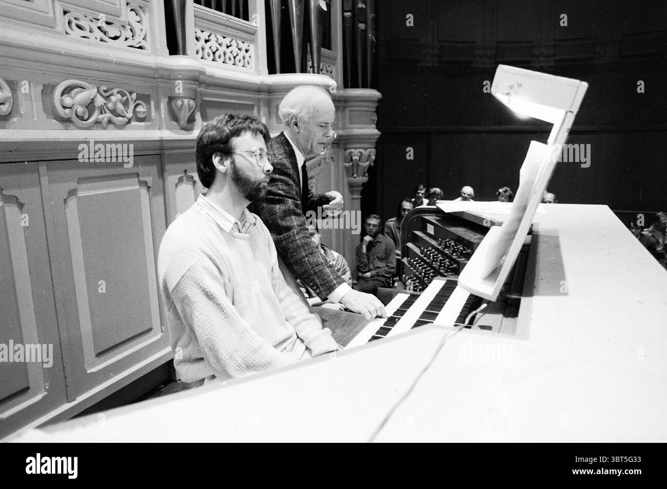 Organ competition master class Concertgebouw Organs and organists, Whizgle News, Dutch Desk, The Netherlands, 1950 - 2000 on 09-07-1988. The image includes these topics. The scene is set inside a grand hall with rich architectural details, characterized by intricate moldings and a large organ as the focal point. The organ has multiple pipes prominently displayed, creating an imposing backdrop, enhanced by soft, diffused lighting that creates a warm atmosphere. Two individuals dominate the foreground. One is seated at the organ, deeply focused on playing, with a serious expression. He has a bea Stock Photo